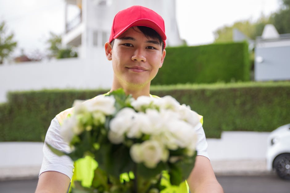 A delivery person wearing a red cap and a reflective yellow safety vest is smiling and extending a bouquet of white roses and green foliage towards the camera outdoors. The bouquet is freshly arranged