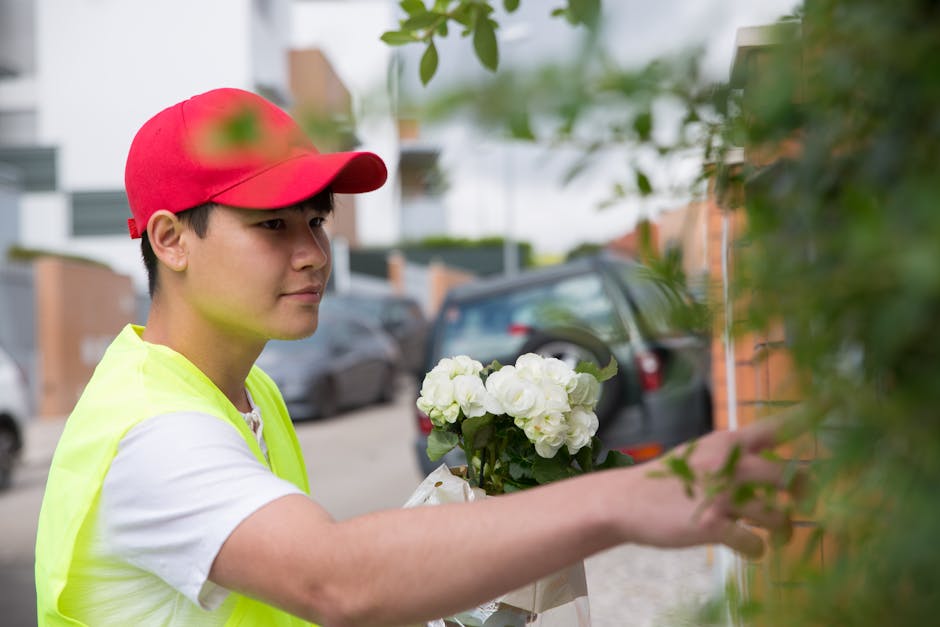 A young delivery person wearing a red cap and a neon yellow high-visibility vest is shown placing a bouquet of fresh white roses and green foliage against a brick wall outdoors. The bouquet is wrapped