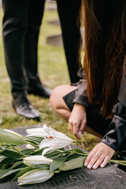 A person in black attire, possibly a woman, is kneeling at a gravesite, gently placing a floral arrangement of white lilies with long green leaves on the gravestone. The lilies have large, open petals
