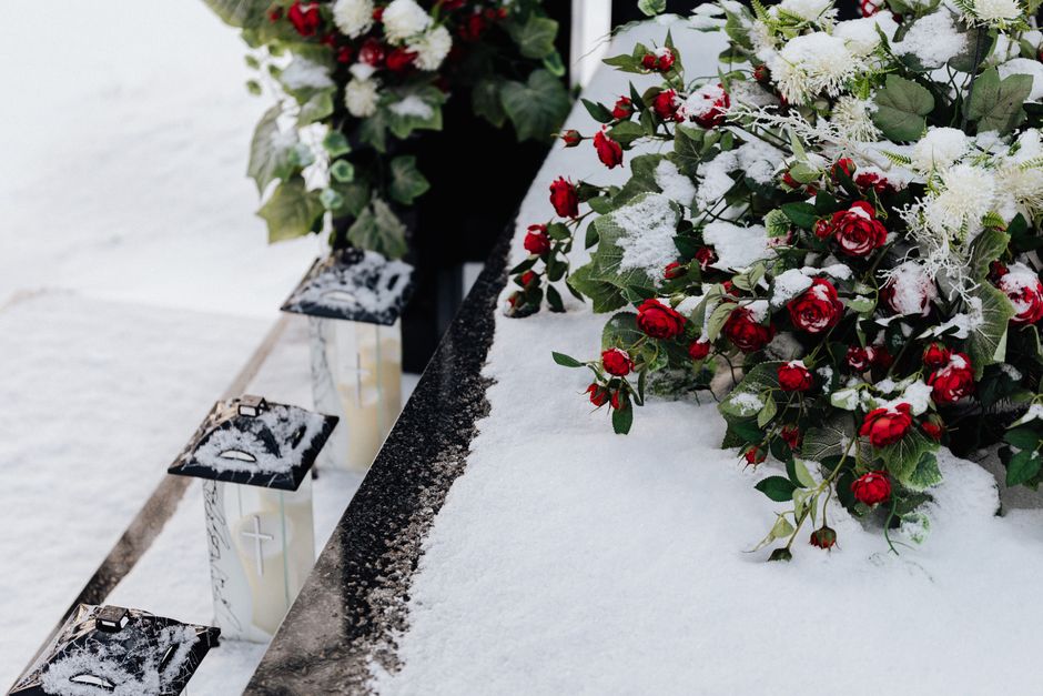 A floral arrangement placed on a snow-covered surface at a cemetery, featuring vibrant red roses and white flowers dusted with snow, with greenery and holly leaves interspersed. The flowers are arrang