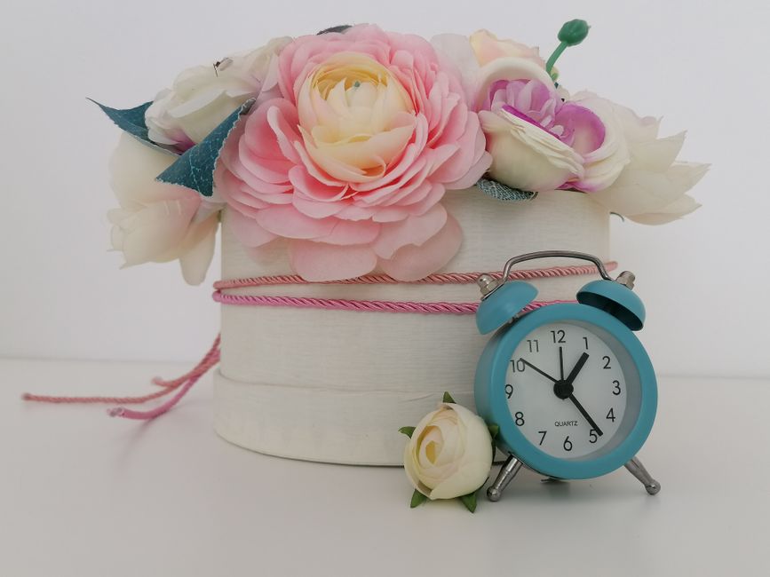 A floral arrangement featuring pink and white roses and peonies in a round white gift box decorated with pink string, with some blue leaves and a pink ribbon. The bouquet is presented on a white surfa