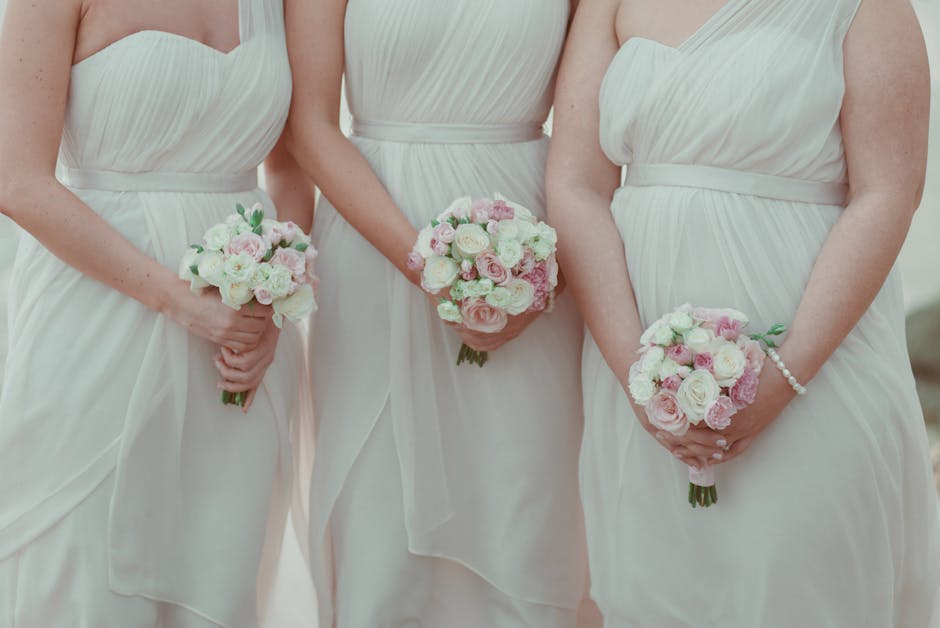 Three women dressed in elegant, strapless white wedding gowns are holding small, round bouquets of fresh flowers. The bouquets comprise white and pale pink roses, complemented by light green foliage,