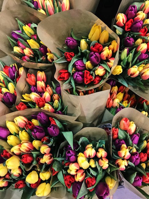 A close-up of multiple bouquets of fresh tulips in vibrant shades of yellow, red, purple, and pink, wrapped in natural brown paper with green foliage accents. The bouquets are arranged in a dense, ove