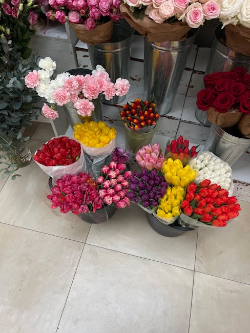 A variety of floral arrangements and bouquets displayed on a tiled floor, including pink roses in metal vases at the back, and smaller bouquets with red, pink, yellow, white, and purple flowers arrang