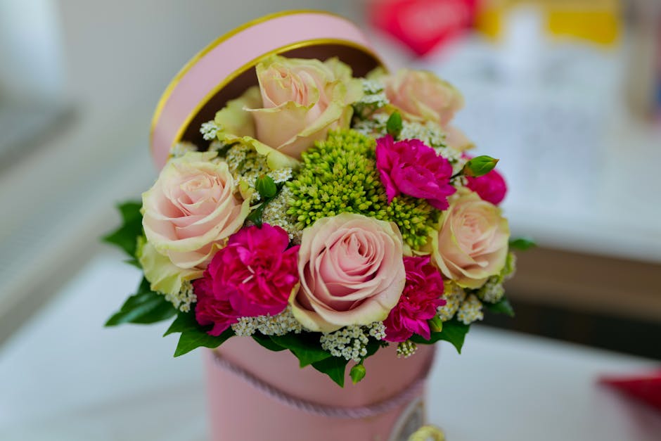A floral arrangement featuring a bouquet of pale pink roses, vibrant magenta carnations, and small clusters of white and green blossoms, arranged in a round, light pink gift box with a ribbon, placed 