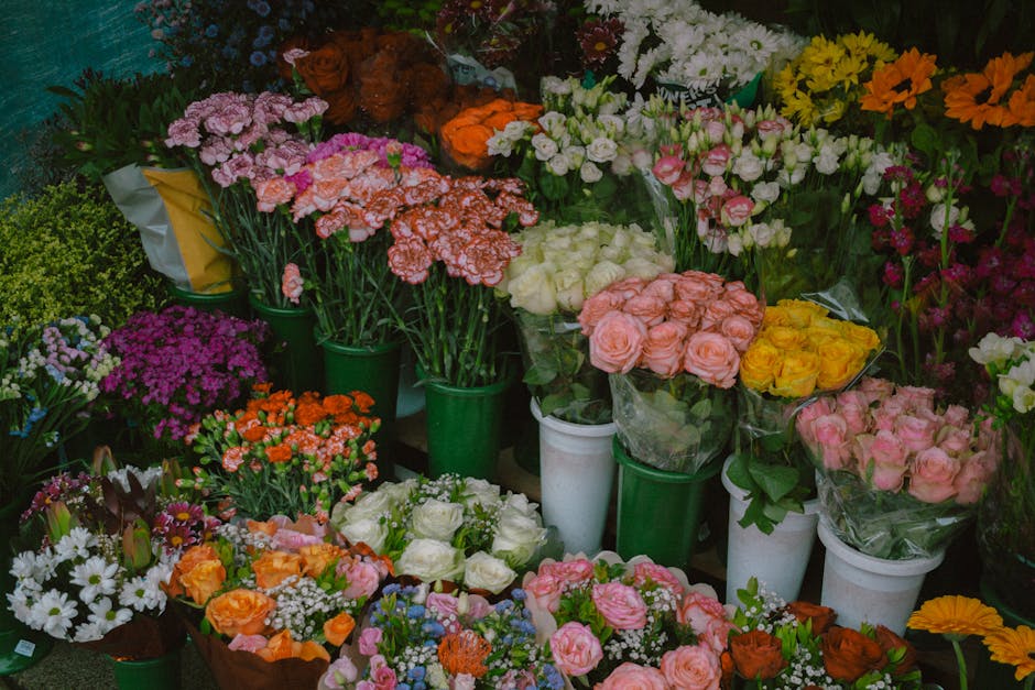 A vibrant display of fresh flowers arranged in various containers at a florist shop, showcasing a wide assortment of flowers including pink roses, white roses, orange carnations, purple chrysanthemums