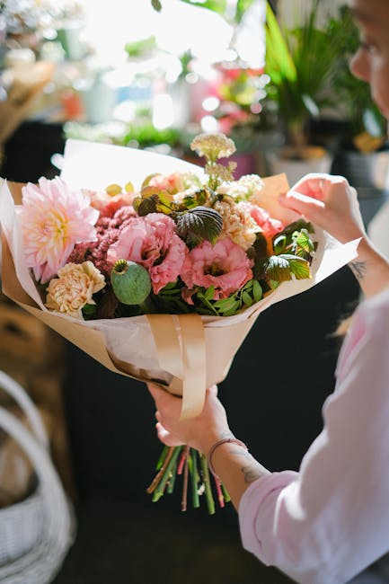 A person holding a large bouquet of fresh flowers wrapped in cream and white paper, featuring predominantly pink peonies, alongside white and purple blossoms and lush green leaves. The bouquet is arra