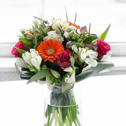 This radiant bouquet, conceived by an expert Ilford florist, is a study in balance, artistry, and local pride. The centre showcases creamy, peach-toned and bright orange gerbera daisies, their petals shimmering in gentle, sunlit waves. Bold magenta roses-tight and richly petalled-offer drama and depth, mirrored by the refined simplicity of white alstroemeria and petite spray roses scattered amongst the ensemble. A scattering of soft pink and delicate white blooms further elevates the arrangement, reminiscent of the tranquil expanses in Valentines Park or the gentle charm of Cranbrook Road in bloom. The base is anchored by broad, glossy green leaves that wrap protectively around the stems, reminiscent of a nurturing embrace within Ilford's welcoming community. Sprigs of smaller, pointed greenery fill out the shape, adding texture and a sense of wildness. Tall, in a clear glass vase, the bouquet's energetic stems contribute to the picture of freshness and organic beauty. Bathed in soft, natural light, this floral arrangement radiates celebration and well-wishing, suitable for milestones, heartfelt thanks, or simply sharing the beauty of expertly crafted flowers from Flower Delivery Ilford.
